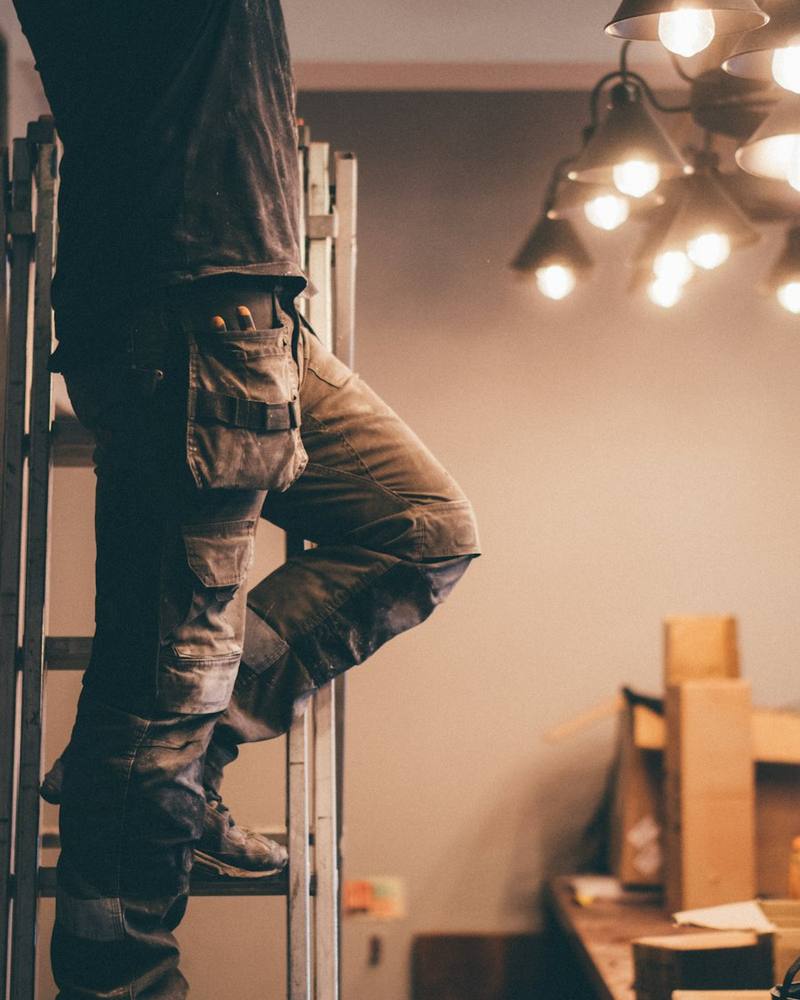 Elektroniker in Arbeitshose mit Werkzeugtaschen auf einer Leiter bei der Montage von Deckenleuchten